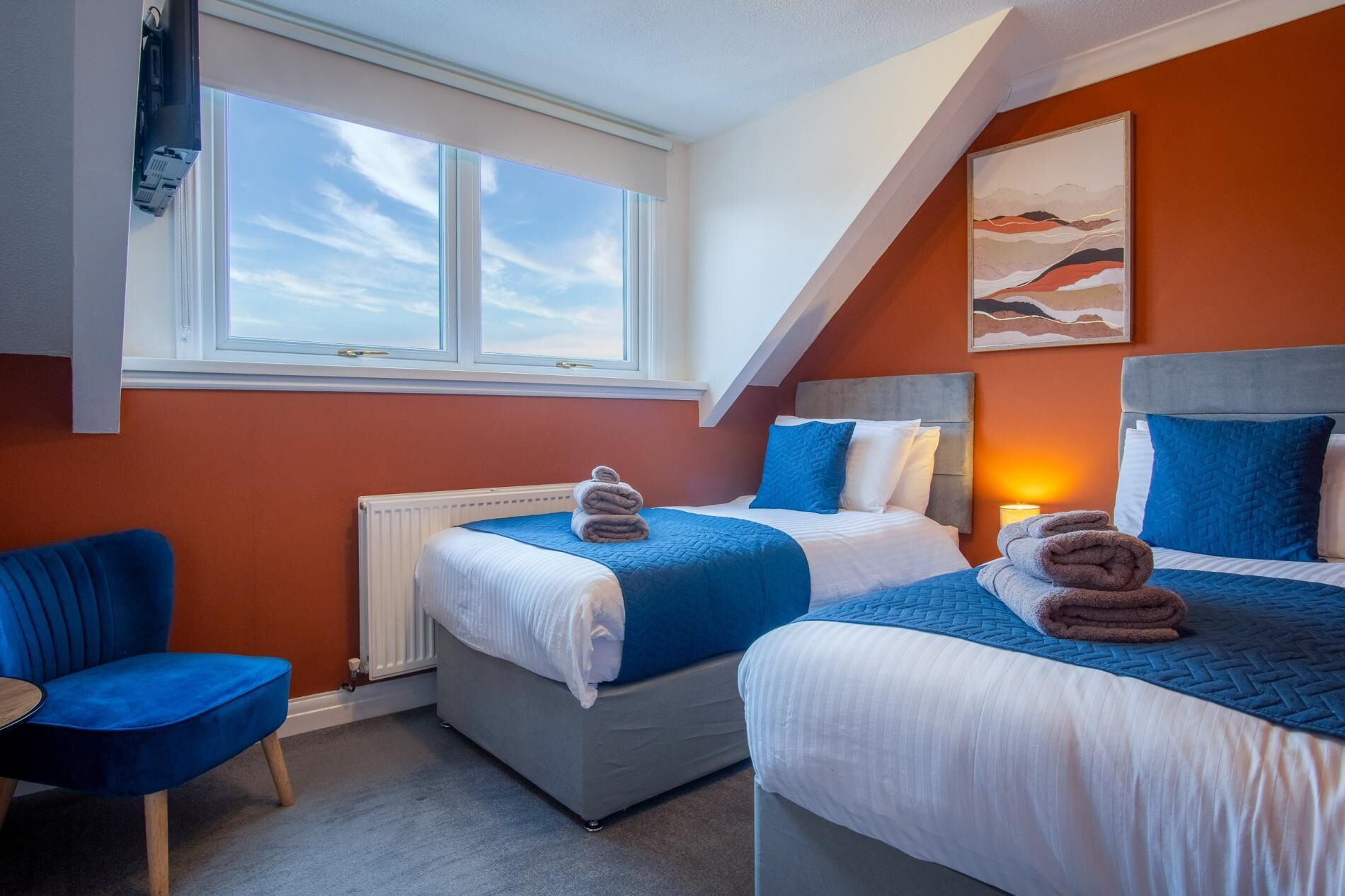 Una camera d'albergo moderna con due letti, una poltrona blu e un'ampia finestra.