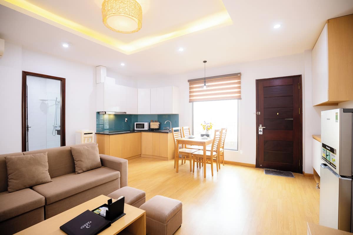 A modern living room with a sofa, a dining table with chairs, a kitchen area with cabinets and appliances, and a wooden floor.