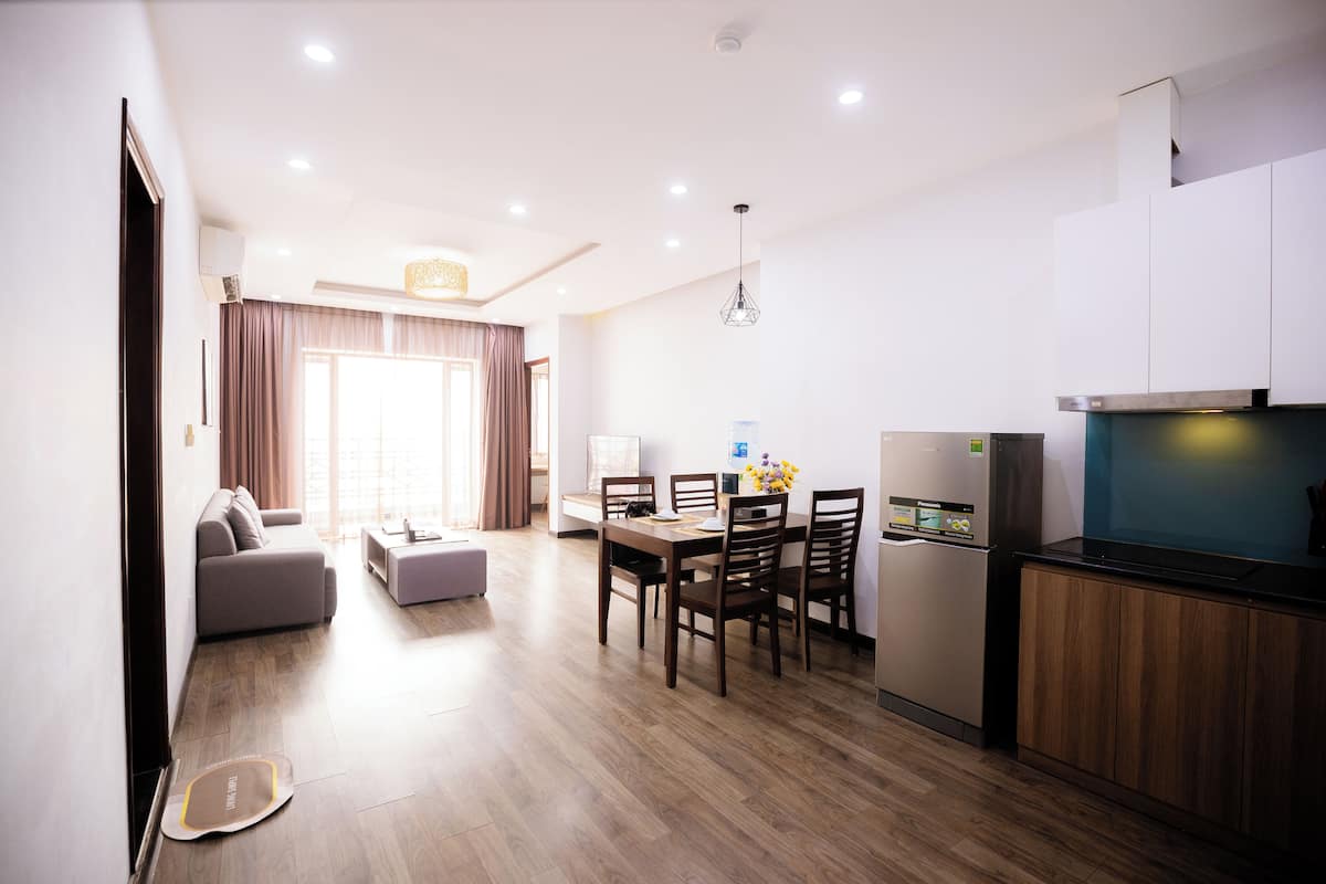 A modern living room with a sofa, dining table, and chairs. There is a kitchen area with a refrigerator and a microwave.