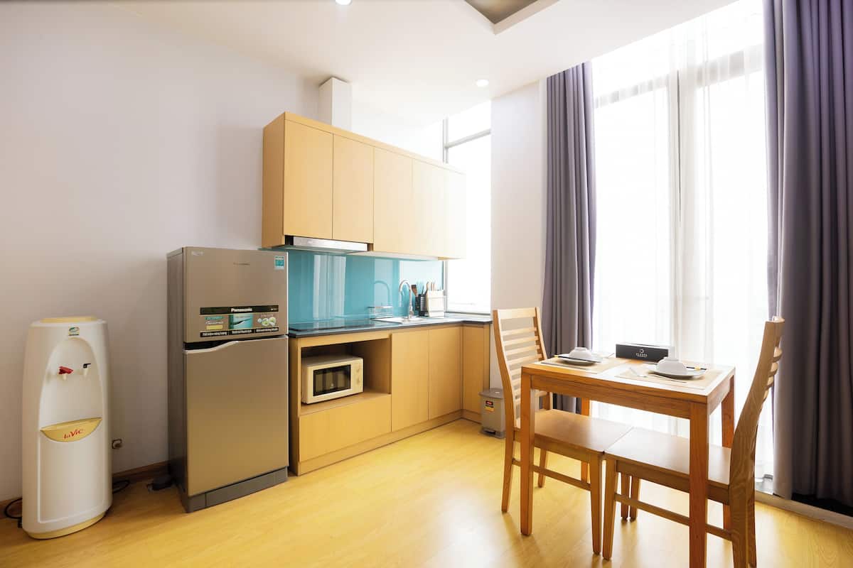 A compact kitchen with a refrigerator, microwave, and a water dispenser. There is a wooden dining table with chairs and a window with curtains.