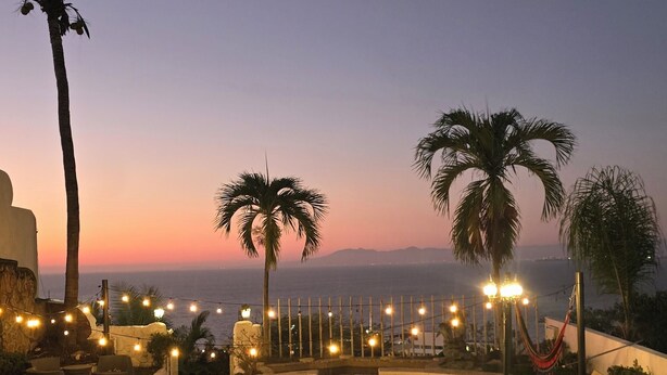Zona de piscina con palmeras, luces colgantes y vista al océano al atardecer.
