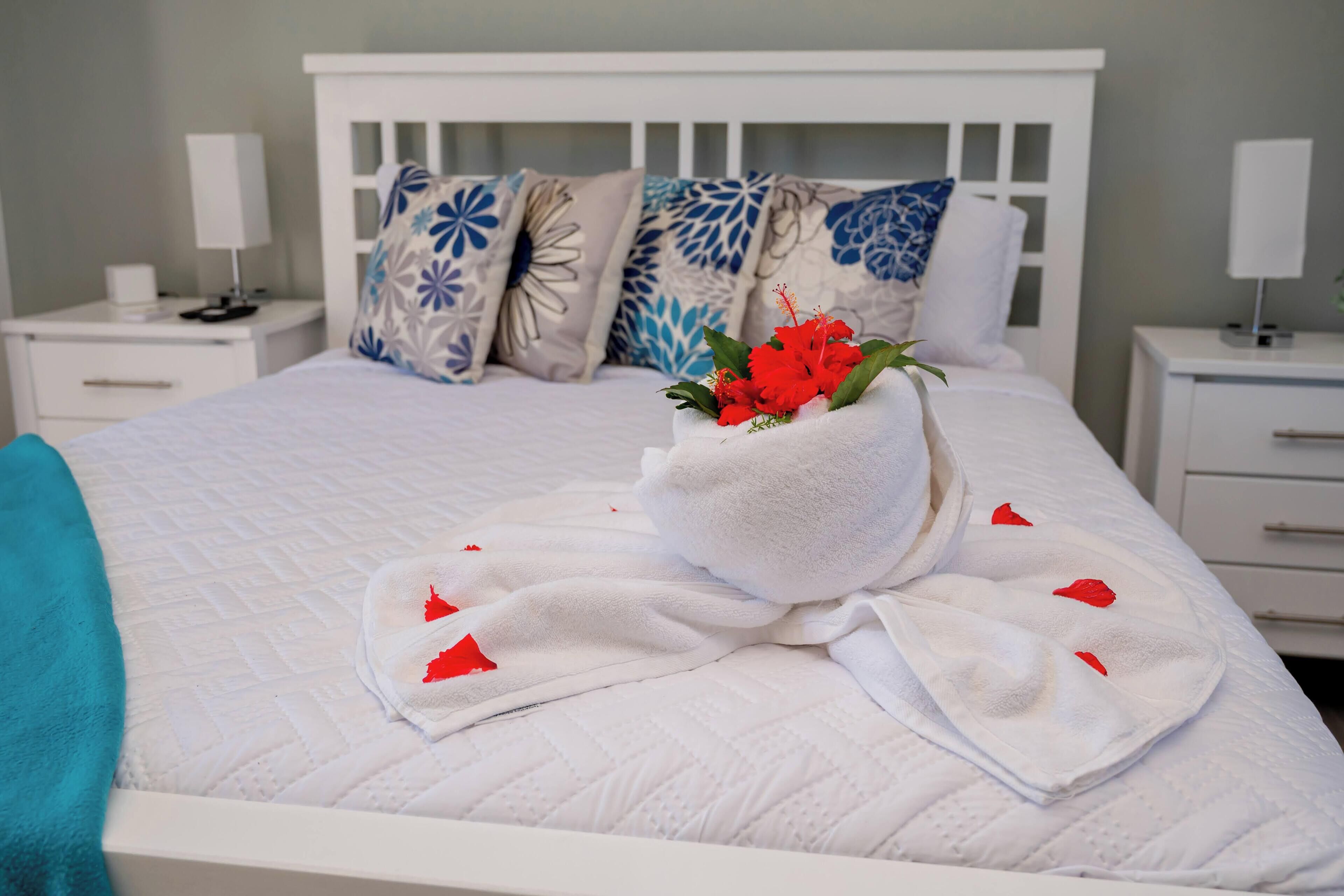 Un lit bien fait, avec une tête de lit blanche, un arrangement décoratif d’oreillers et une serviette blanche ornée de fleurs rouges posée sur le lit.