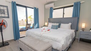 A bedroom with a large bed, a bedside table, a nightstand, a fan, and a window with curtains.