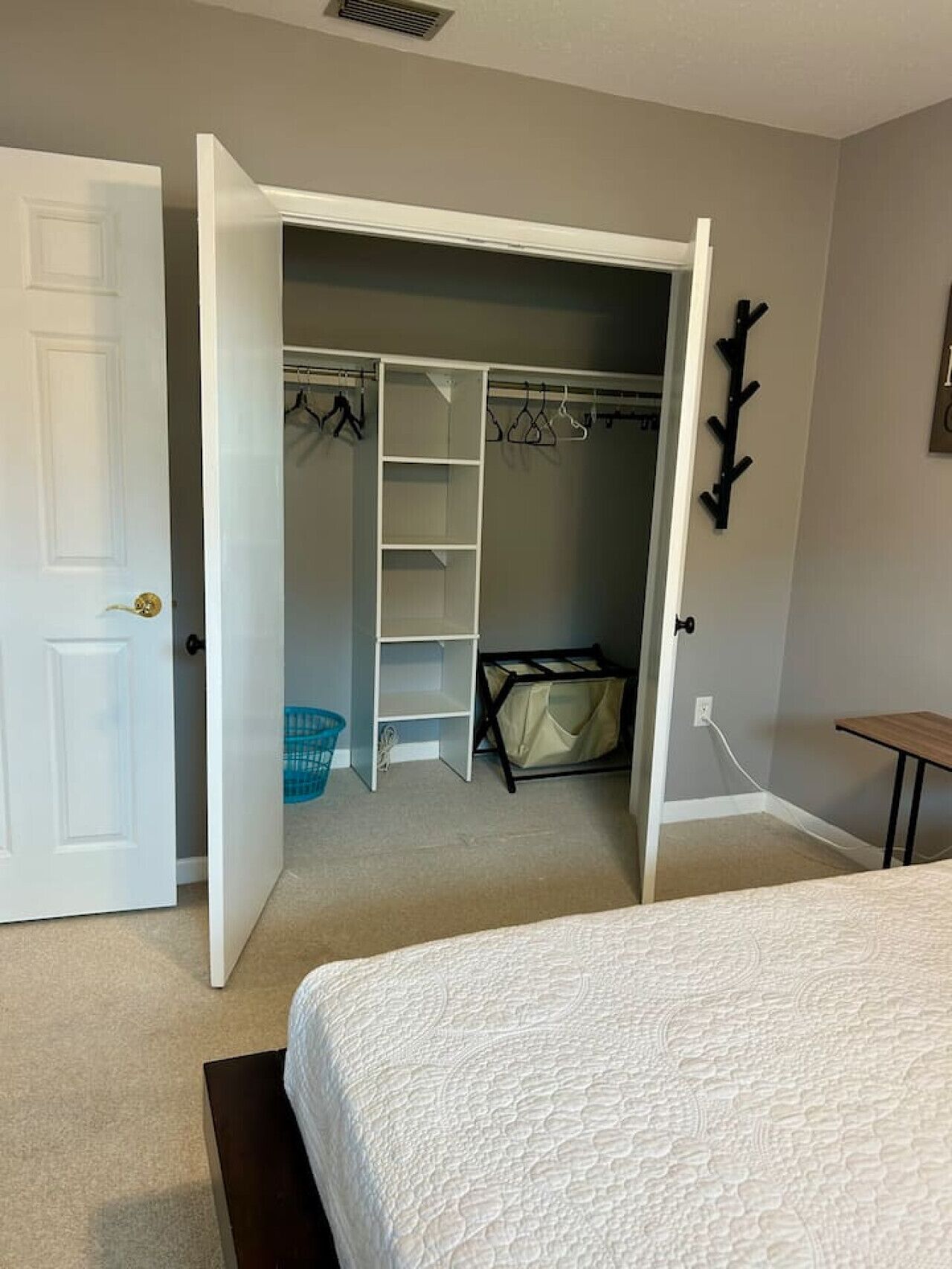 A bedroom with a bed, an open closet with shelves and hanging space, a blue laundry basket, and a small table.