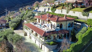 Un quartier résidentiel situé sur une colline, avec une maison remarquable qui se distingue par son toit recouvert de tuiles rouges, un escalier en pierre et une terrasse aménagée avec du mobilier d’extérieur.