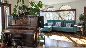 Un salon avec un grand piano, un canapé de couleur turquoise et une grande fenêtre donnant sur la forêt.