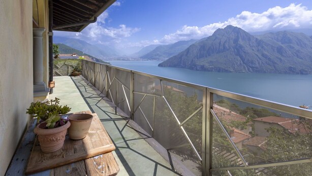 Un balcon avec vue sur un lac et des montagnes, une table en bois agrémentée de plantes en pot et une rambarde en métal.