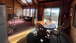 A cozy cabin interior with a wooden dining table, chairs, a sofa, and a large window offering a view of the outdoors. - Ocean View, Cozy 2-bedroom cabin in tranquil Gualala with WiFi and huge firepit (gualala)