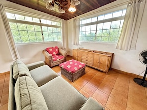A room with a sofa, a wicker chair with a red cushion, a wooden cabinet, and large windows with curtains.