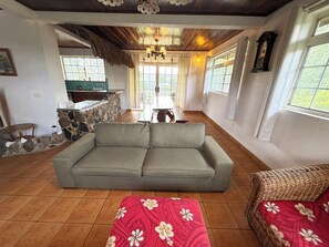 A cozy living room with a sofa, a red patterned ottoman, a wicker chair, a stone wall, a kitchen area, and large windows.