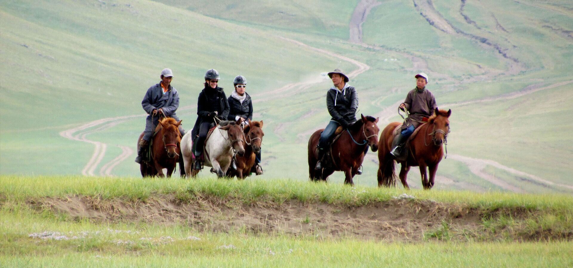 Un grupo de personas montando a caballo en una colina cubierta de hierba, con un camino sinuoso al fondo.