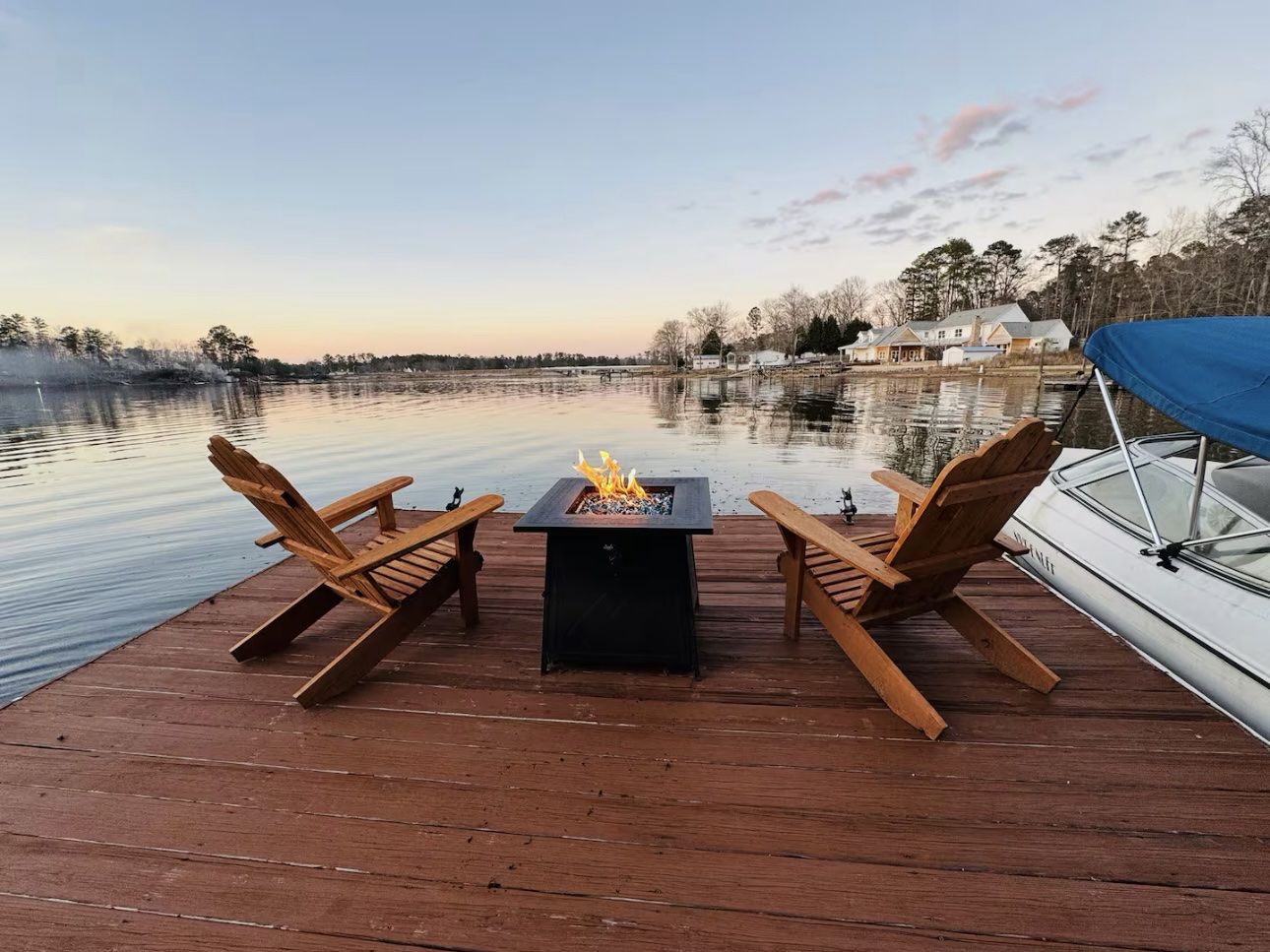 A wooden deck with a fire pit table and two chairs facing a calm lake.
