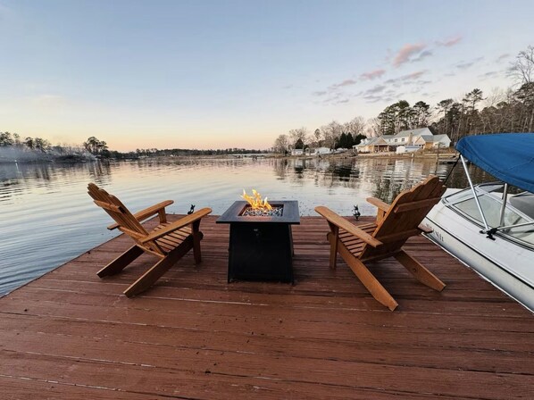 Una terraza de madera con una mesa para fuego y dos sillas frente a un lago tranquilo.