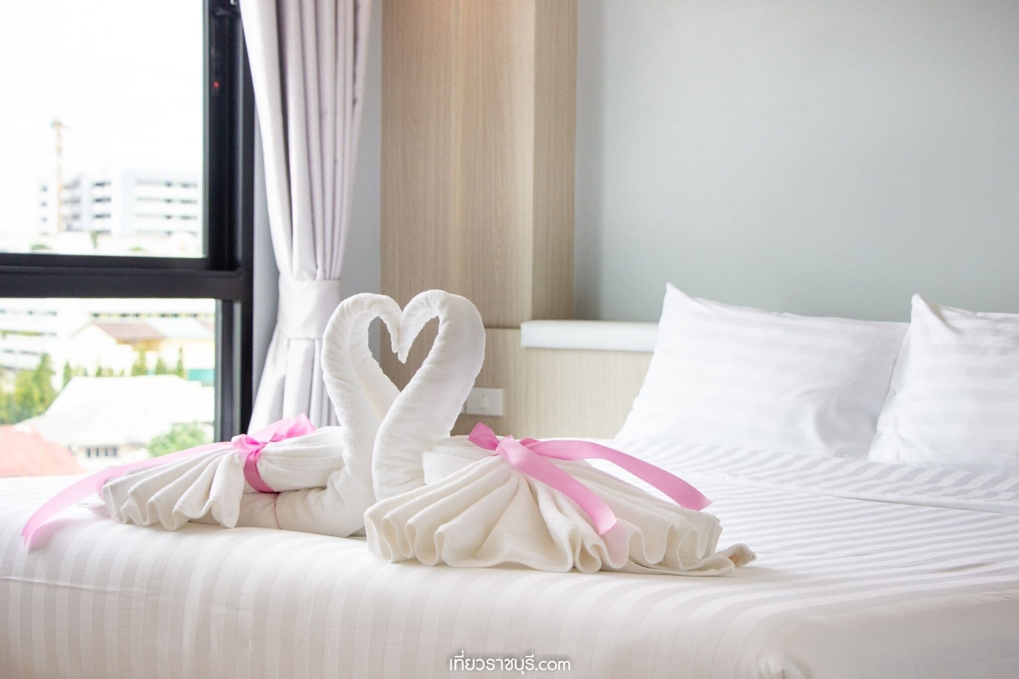 A neatly made bed with white pillows and a heart-shaped towel decoration tied with a pink ribbon.