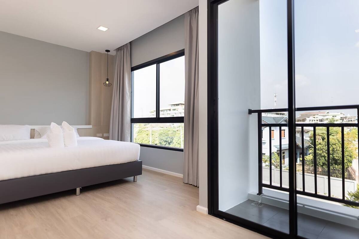 A modern bedroom with a large bed, a view of the city through large windows, and a balcony with a railing.