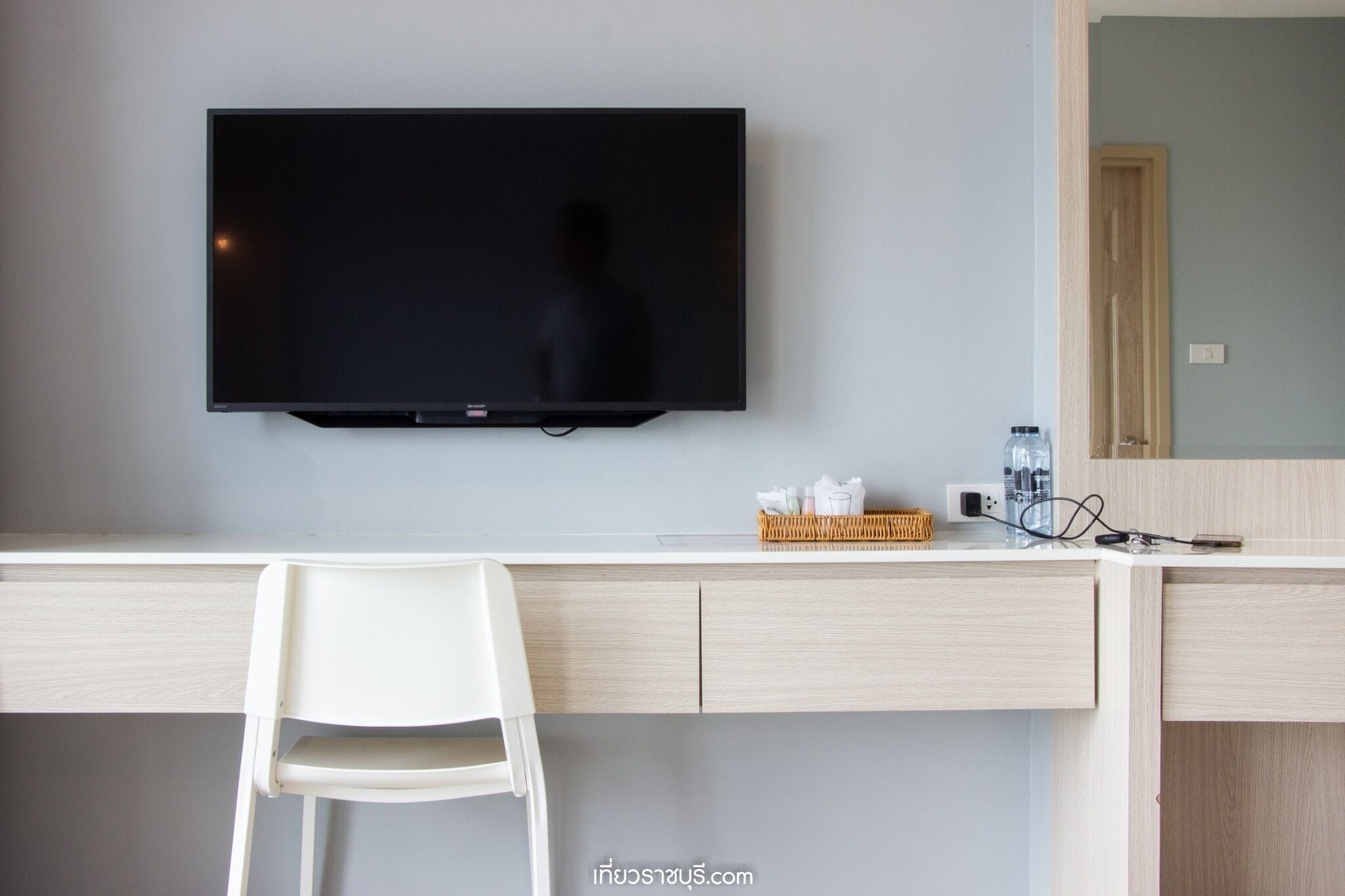 Habitación de hotel moderna con televisor de pantalla plana en la pared, un escritorio blanco con una silla y una pequeña cesta de mimbre sobre el escritorio.