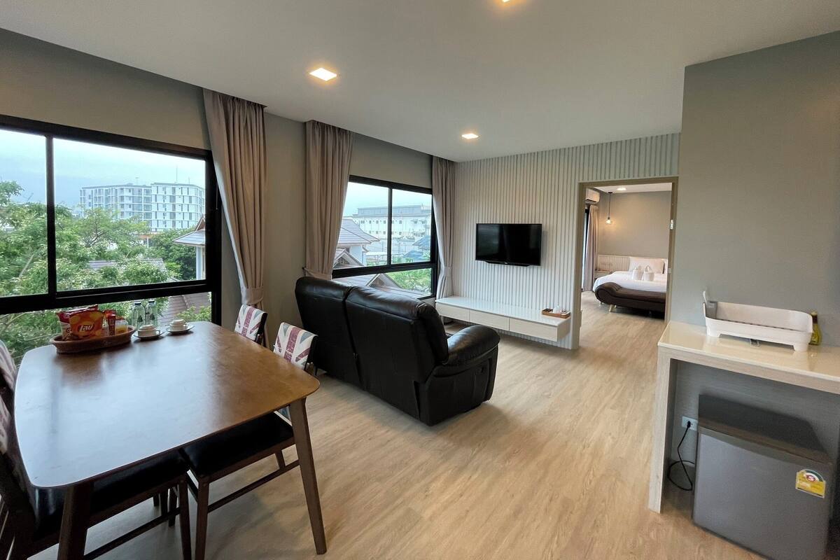 A modern hotel room with a dining area, a sofa, a TV mounted on the wall, and a view of the cityscape.