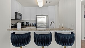 A modern kitchen with white cabinets, a stainless steel refrigerator, a granite countertop, and three dark blue bar stools. - Oceanfront Oasis: Serene 1bd Retreat in MDR/Venice (Marina del Rey)