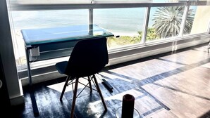 A room with a large window overlooking the ocean, a desk with a chair, and a small table with a candle. - SeafrontApartment-Sleeps4-Pool-Parking-Garden (Porlamar)
