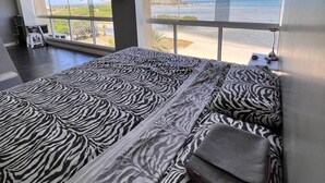A bedroom with a zebra-patterned bedspread, a large window with a view of the sea and a distant cityscape, and a flat-screen TV mounted on the wall. - SeafrontApartment-Sleeps4-Pool-Parking-Garden (Porlamar)