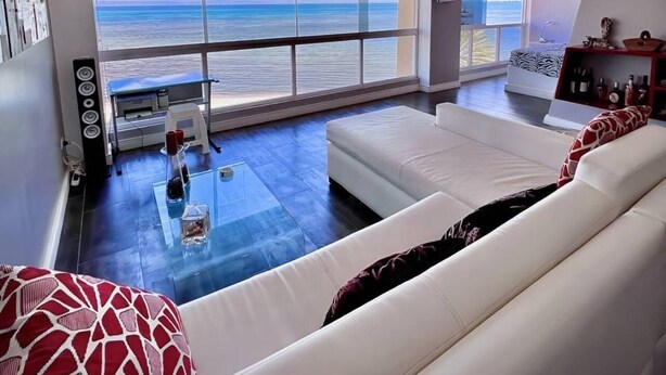 A modern living room with a large window overlooking the ocean, a white sofa, and a glass coffee table. - SeafrontApartment-Sleeps4-Pool-Parking-Garden (Porlamar)