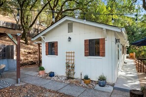 Un petit bâtiment blanc avec des volets en bois, une échelle en bois et des plantes en pot.