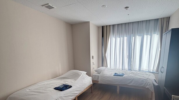 A small, clean room with two single beds, a wooden floor, a ceiling with a light fixture, and a window with curtains. - SakuraMandarin (Sakura)