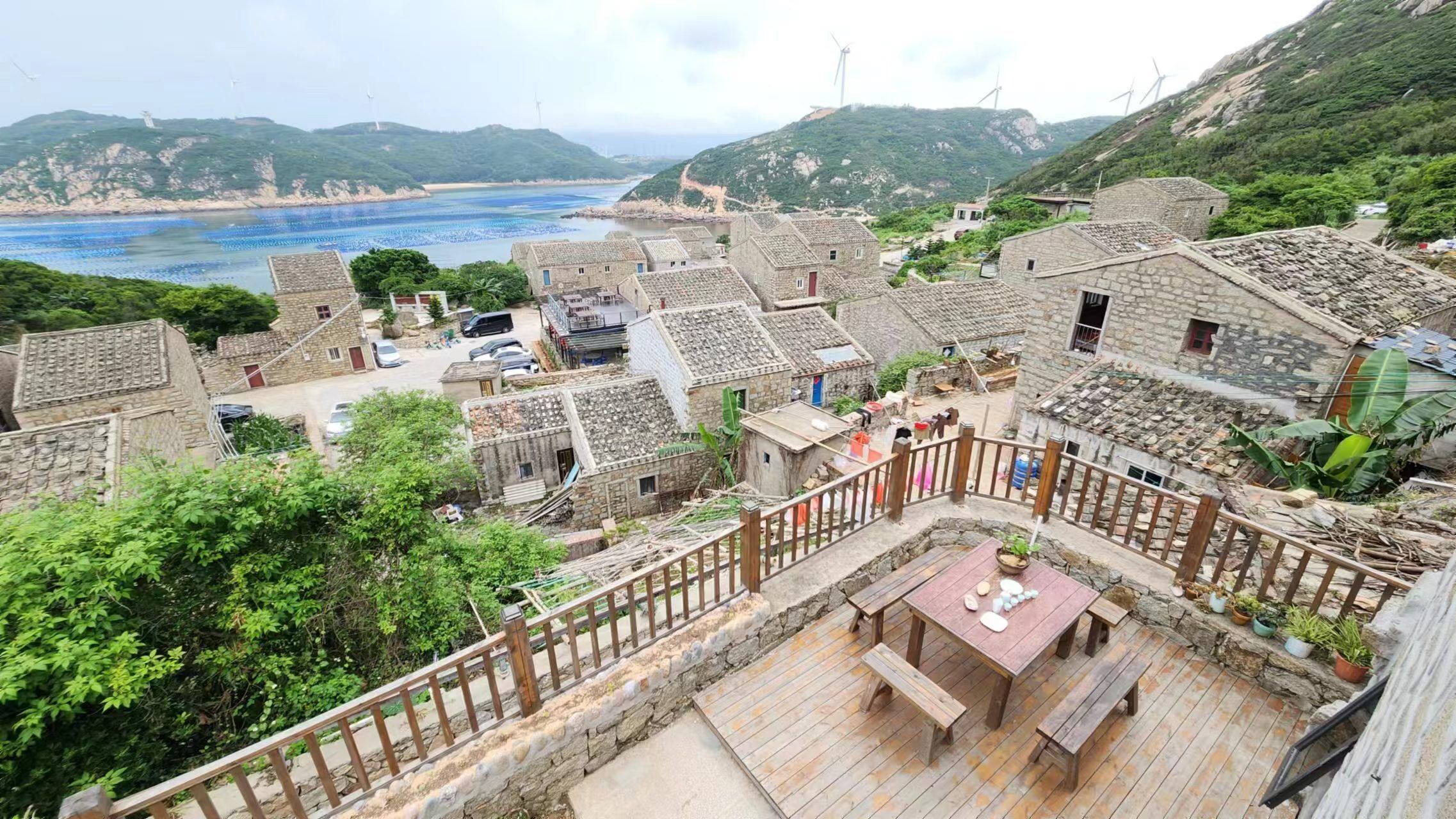 A traditional village with stone buildings, a wooden deck with seating, and a view of the sea and wind turbines in the background.