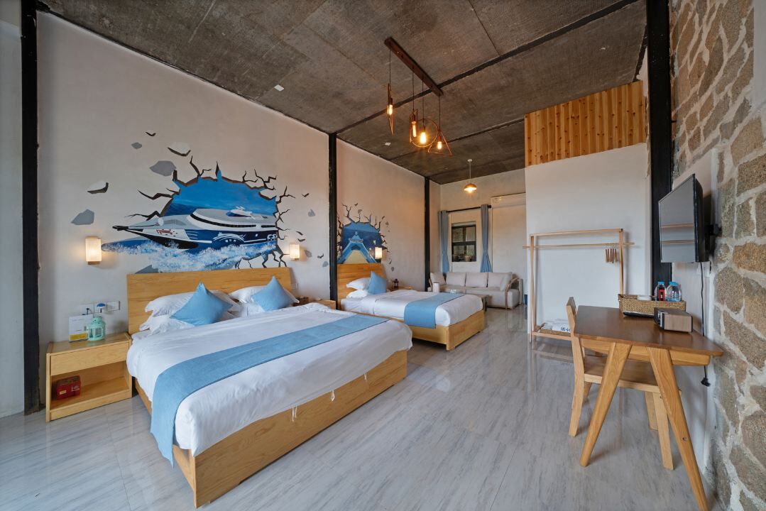 A modern hotel room with two beds, a wooden desk, and a stone wall.