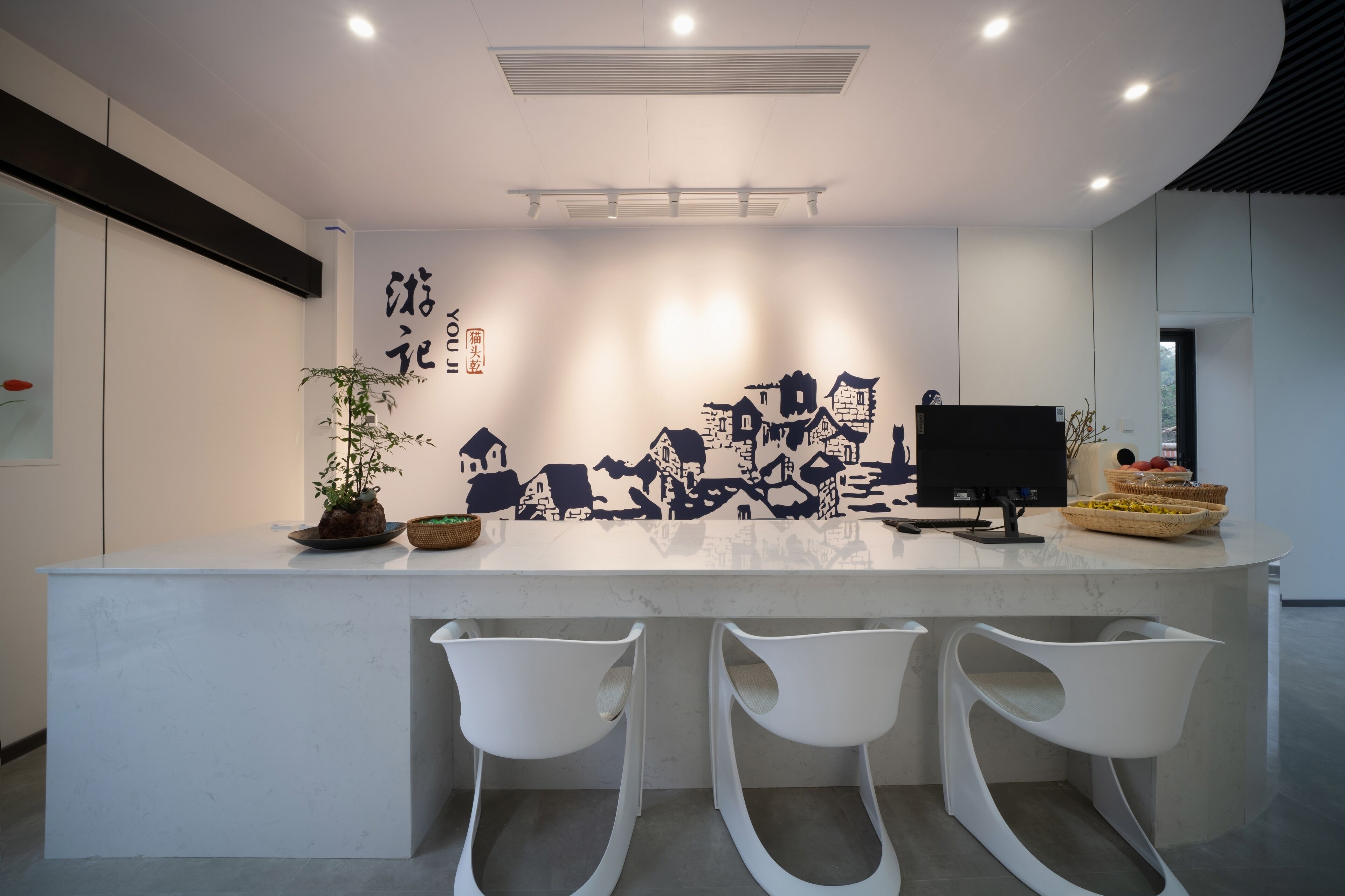 A modern kitchen with a white countertop, three white bar stools, a black monitor, and a decorative wall with black and white patterns.