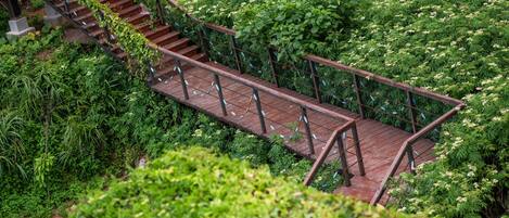 Un puente de madera con barandillas metálicas que une dos zonas elevadas, rodeado de abundante follaje y plantas en flor.