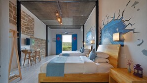Une chambre avec un grand lit, une tête de lit en bois et une vue sur l’extérieur. On y trouve un bureau avec une chaise et une petite table sur laquelle est posé un vase de fleurs.