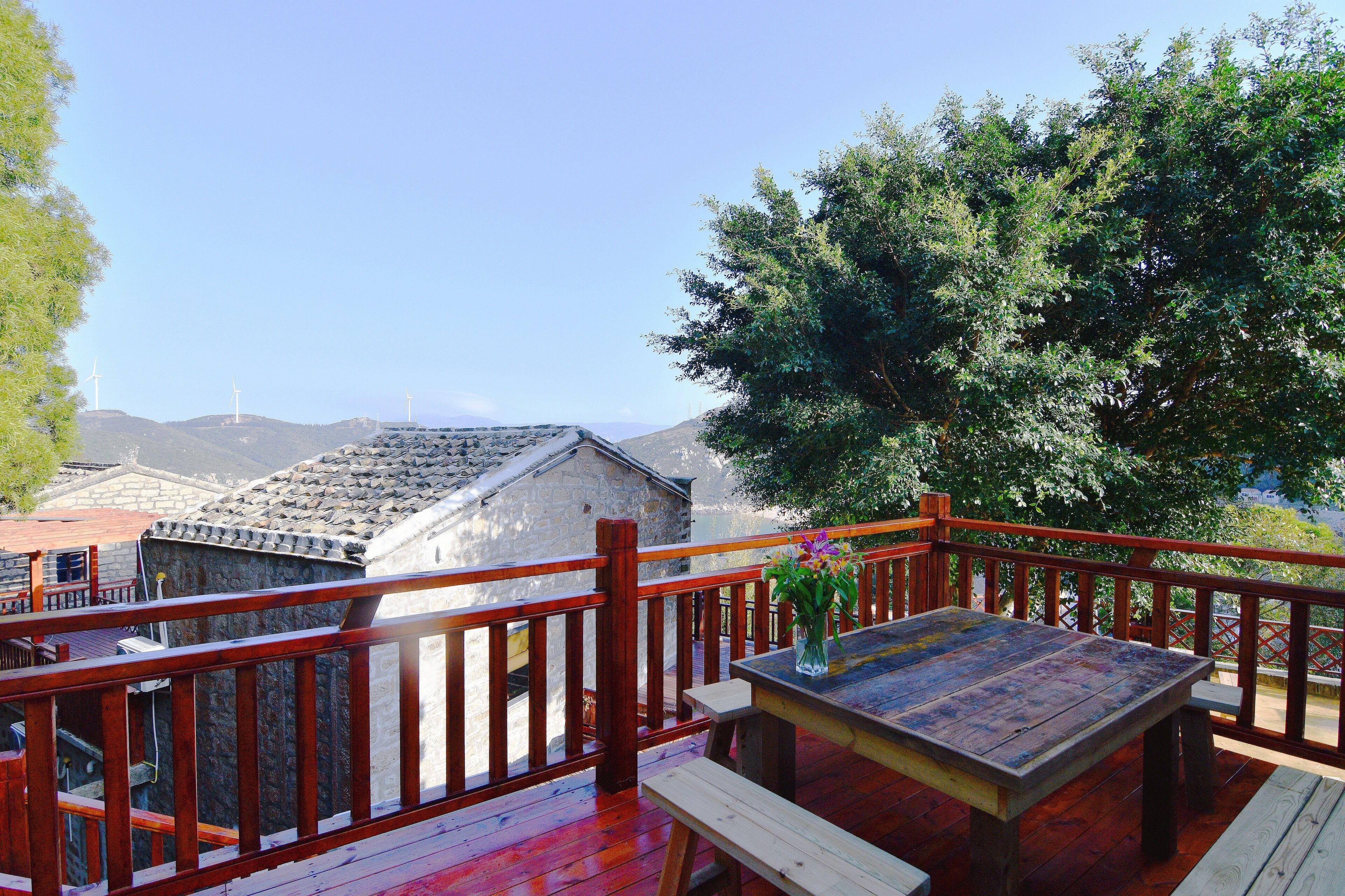 Una terraza de madera con una mesa y bancos, con vistas a un paisaje montañoso con aerogeneradores.