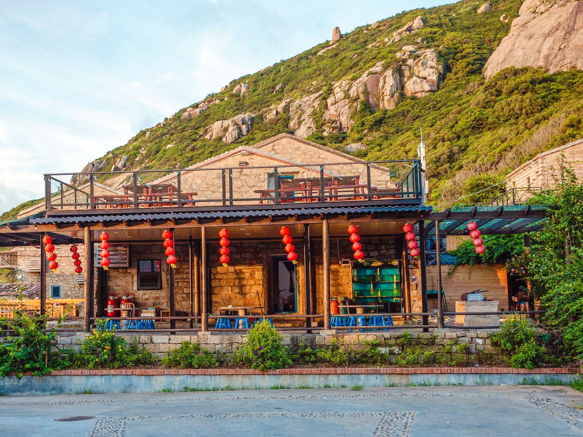 A rustic stone building with a balcony and red lanterns, surrounded by greenery and hills.