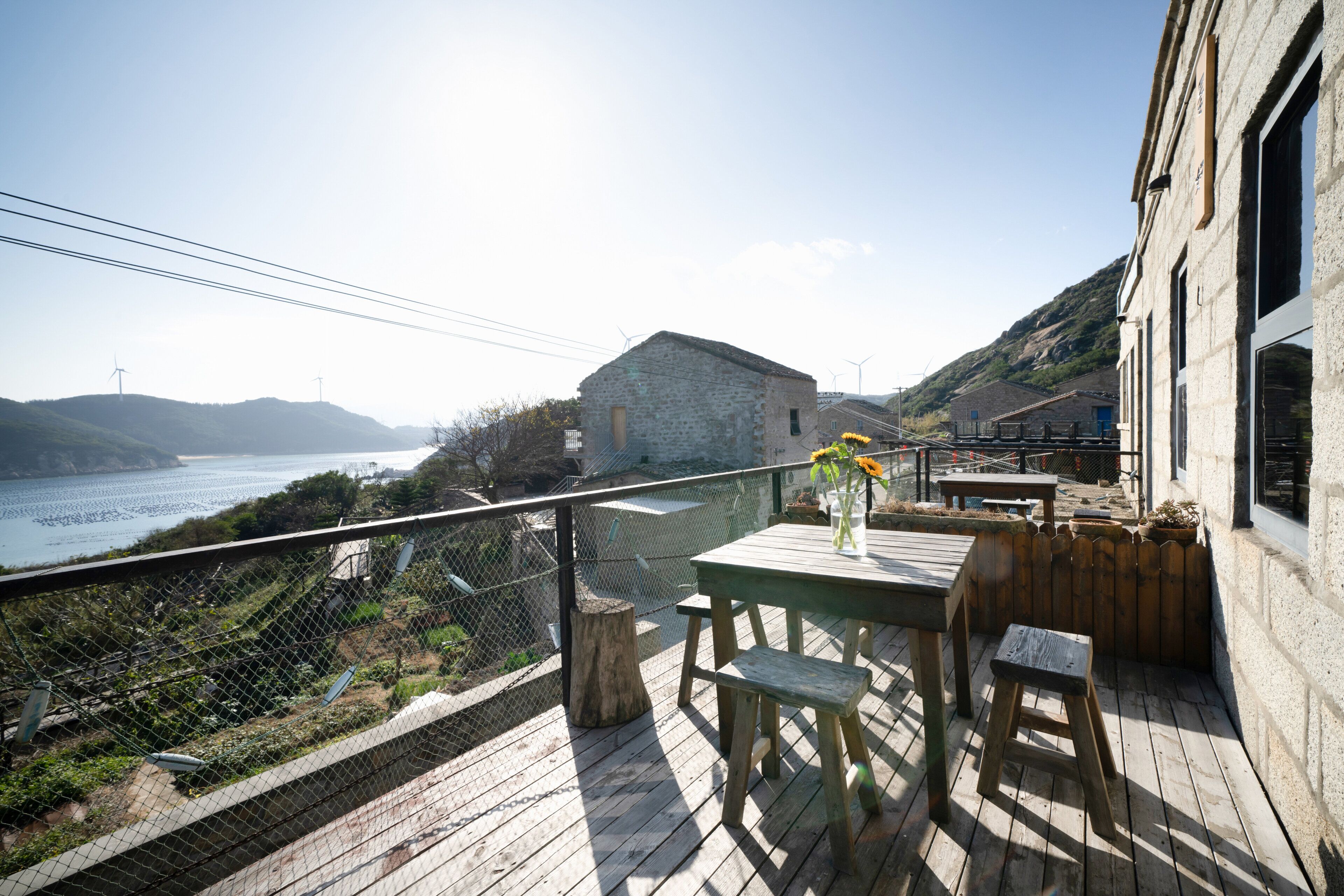 Une terrasse en bois avec une table et un banc, donnant sur une zone côtière où se trouvent des éoliennes et un bâtiment en pierre.