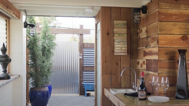 A narrow corridor leading to a wooden kitchen area with a sink, wine bottle, and glasses on the counter. A large potted plant is visible in the background.
