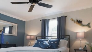 A bedroom with a bed, a ceiling fan, a nightstand with a lamp, a mirror, and a window with curtains.