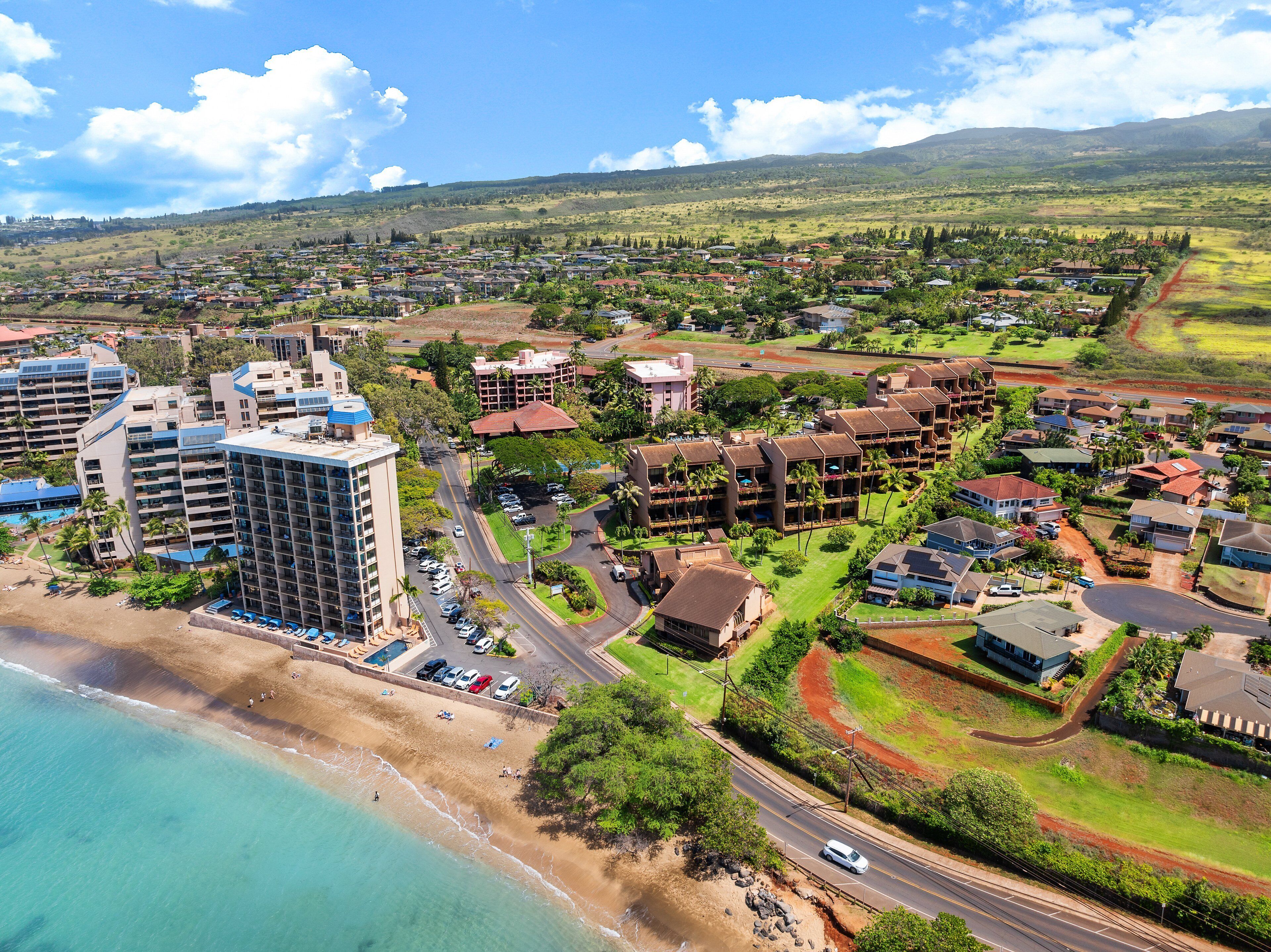 An aerial view of a coastal resort area with beachfront properties, a main road, and a hotel with multiple floors and balconies.