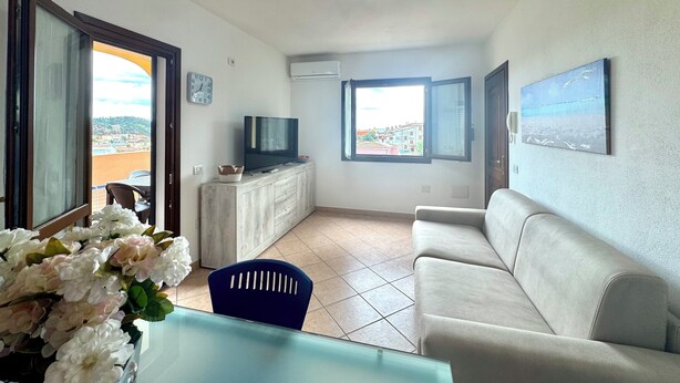 Olohuoneessa on sohva, kaapilla oleva televisio, ruokapöytä tuoleineen, ikkunasta avautuva näkymä ja seinällä oleva kello.