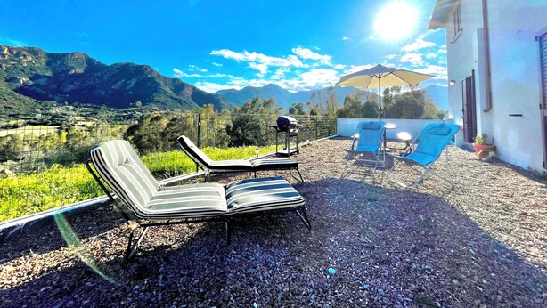 Un patio con dos sillones reclinables a rayas, una mesa, una parrilla y una sombrilla, con vistas a un paisaje montañoso.