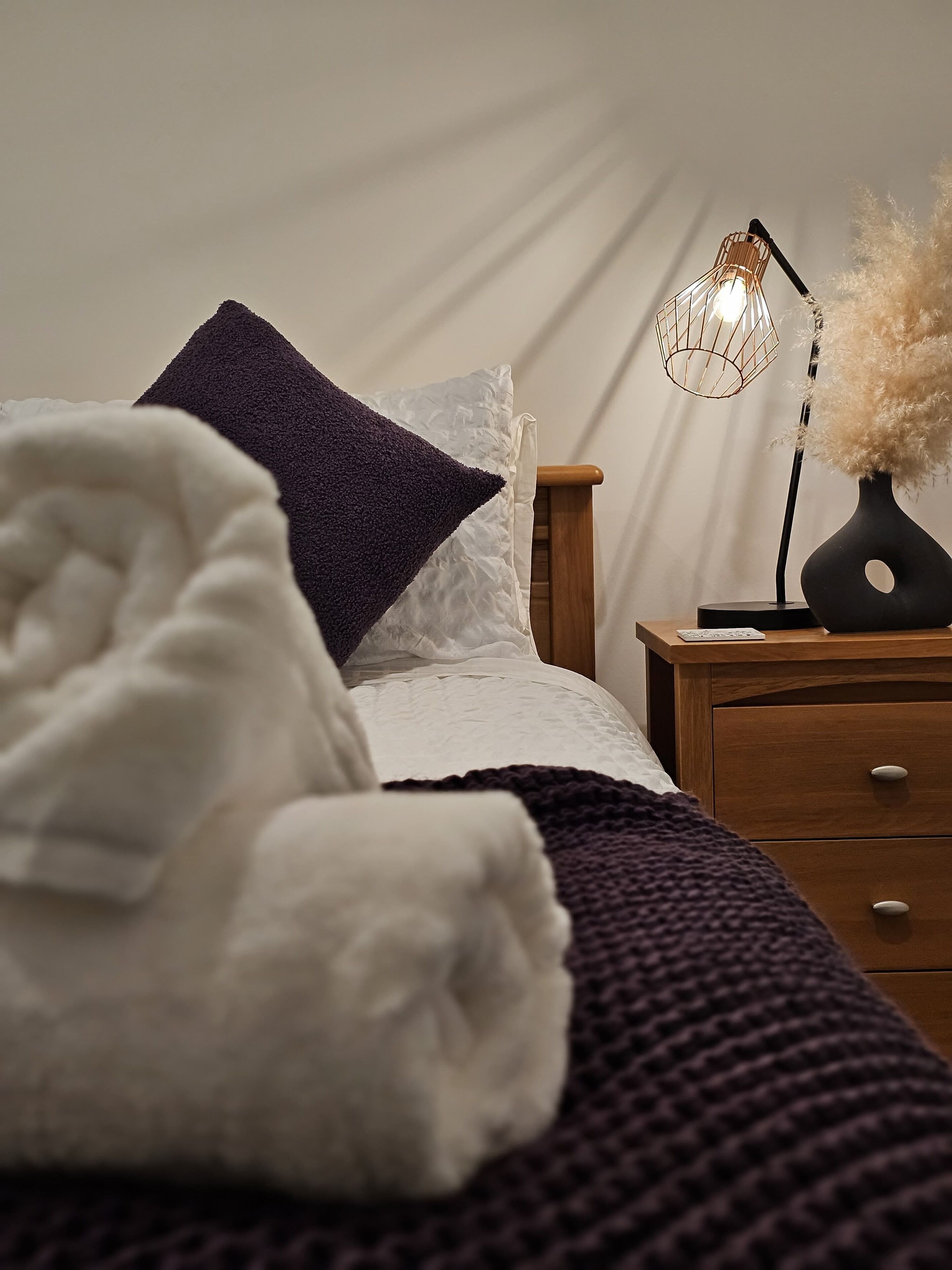 Un letto ben rifatto con coperta bianca e cuscino blu scuro, un comodino in legno con una lampada, e una parete bianca sullo sfondo.