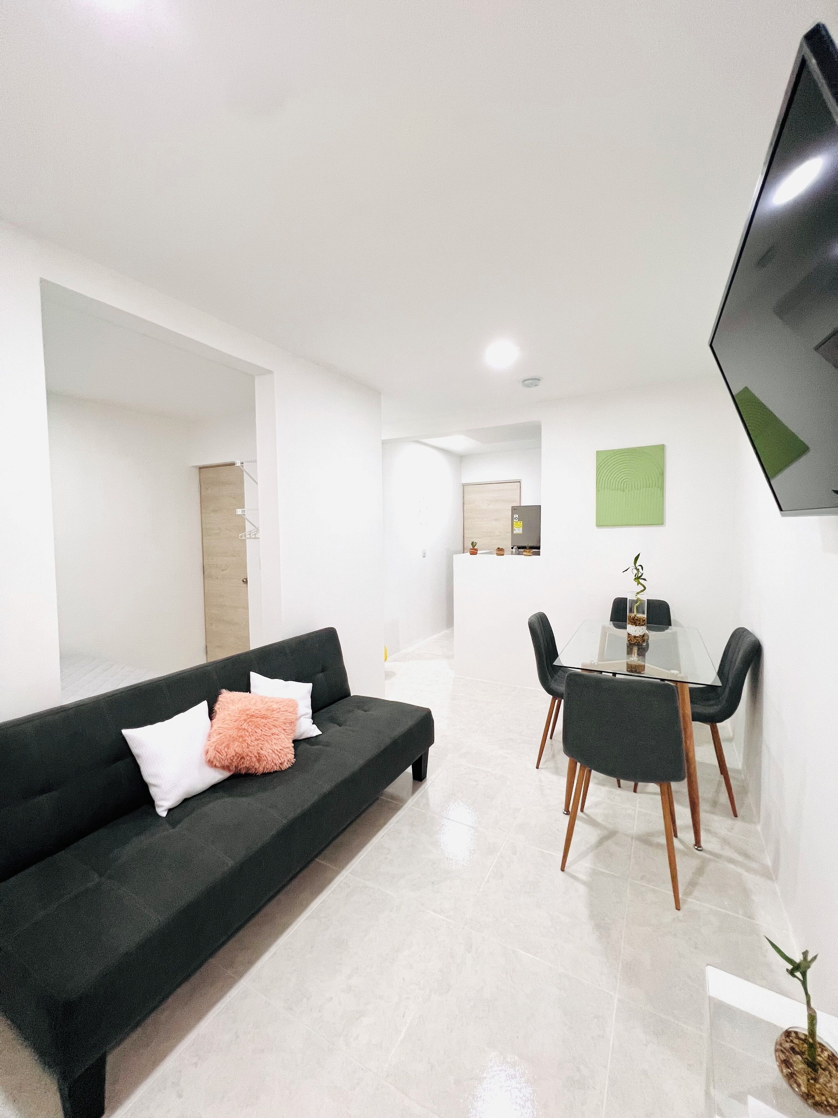 A modern living room with a dark green sofa, a glass dining table, and a small kitchen area in the background.