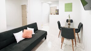 Un soggiorno moderno con un divano verde scuro, un tavolo da pranzo in vetro e una piccola zona cucina sullo sfondo.