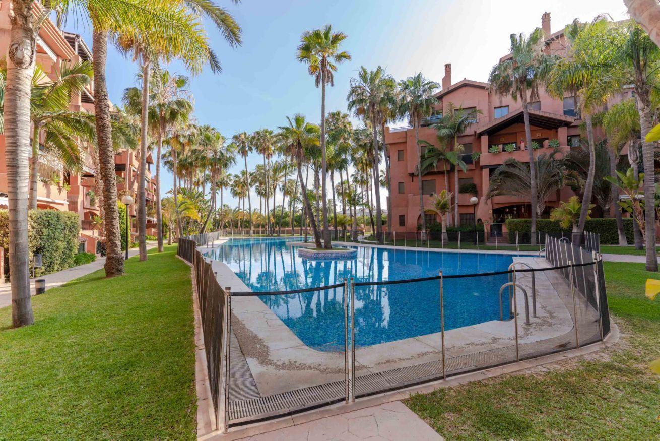 A swimming pool surrounded by palm trees and a red building with balconies.