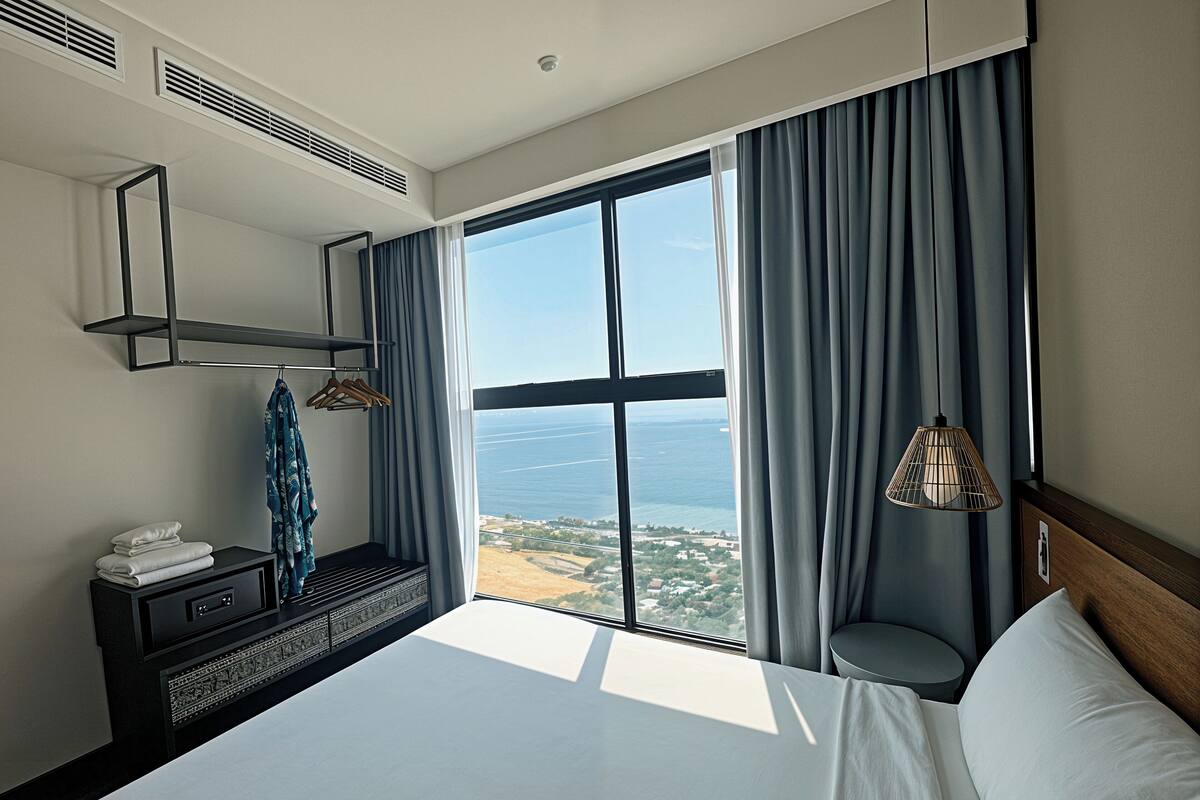 A hotel room with a large window, a bed, a bedside table, a mirror, and a hanging light fixture.