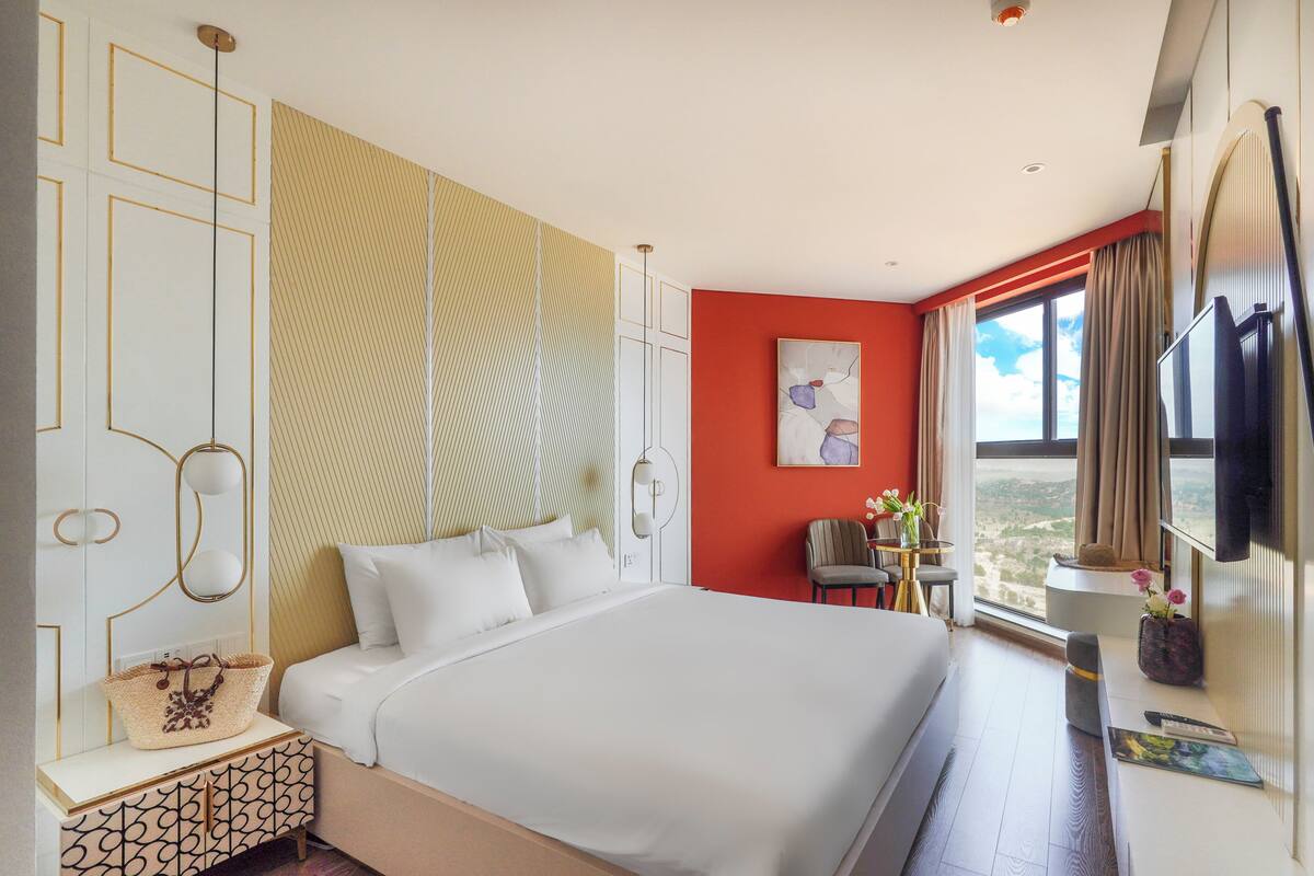 A modern hotel room with a large bed, a desk with a chair, a small table with a vase, and a window with a view.