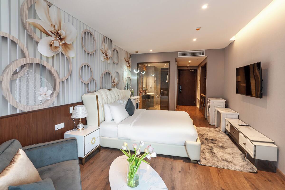 A modern hotel room with a large bed, a bedside table, a chair, a small table with a vase, and a TV mounted on the wall.
