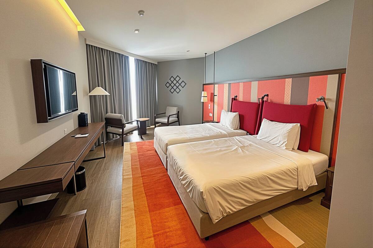 A hotel room with two beds, a desk, a chair, a TV, and a window with curtains.