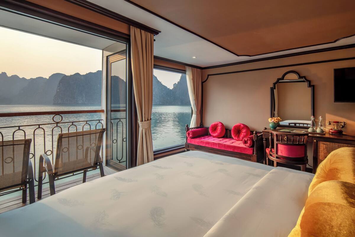 Quarto de hotel com uma cama grande, um sofá, uma poltrona e uma televisão. O quarto tem vista para a água e montanhas.