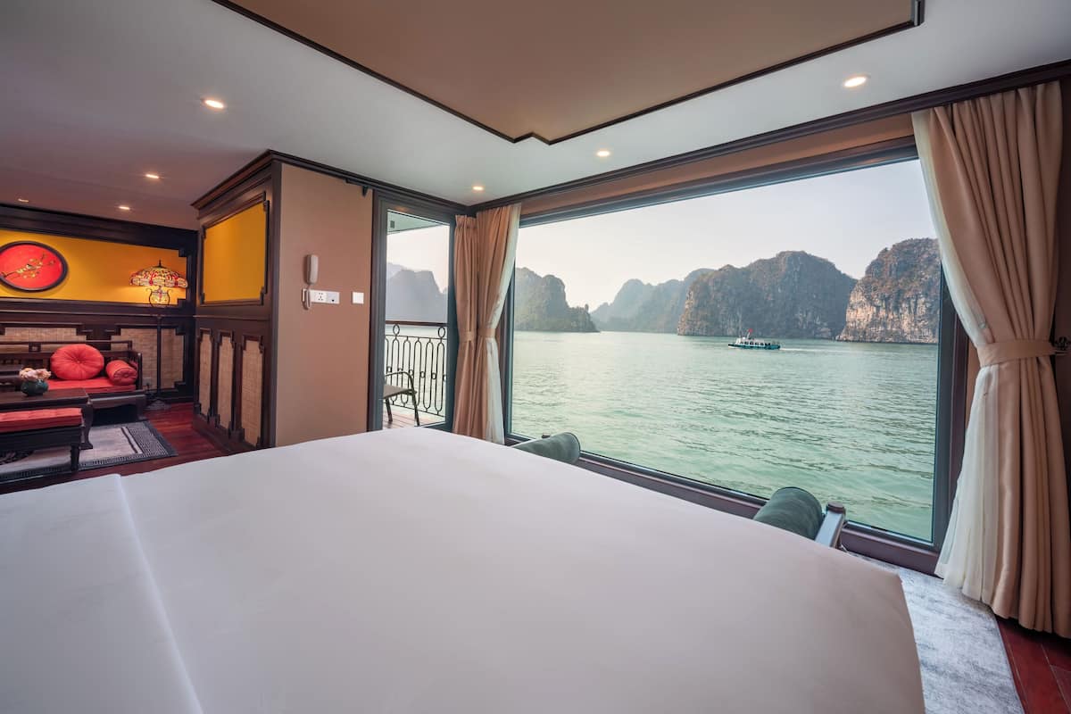 Quarto de hotel com uma cama grande, um espaço de estar pequeno com um almofada vermelha, um telefone fixado na parede e vista para um barco na água.